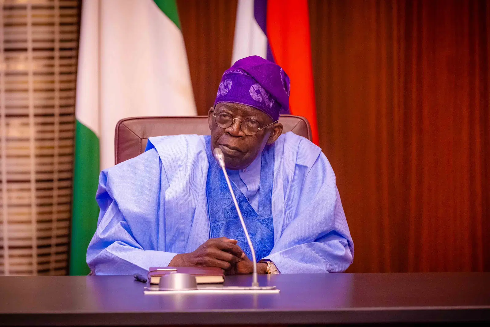 President Bola Tinubu seated at a desk with a microphone in front of Nigerian flags