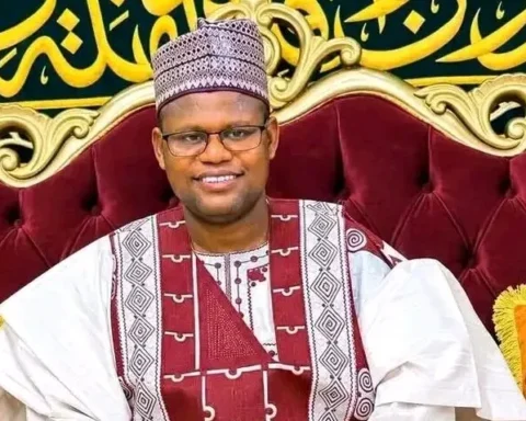 Sheikh Yahaya Masussuka, the Katsina cleric, in a white and maroon traditional outfit and glasses, smiling on a red sofa