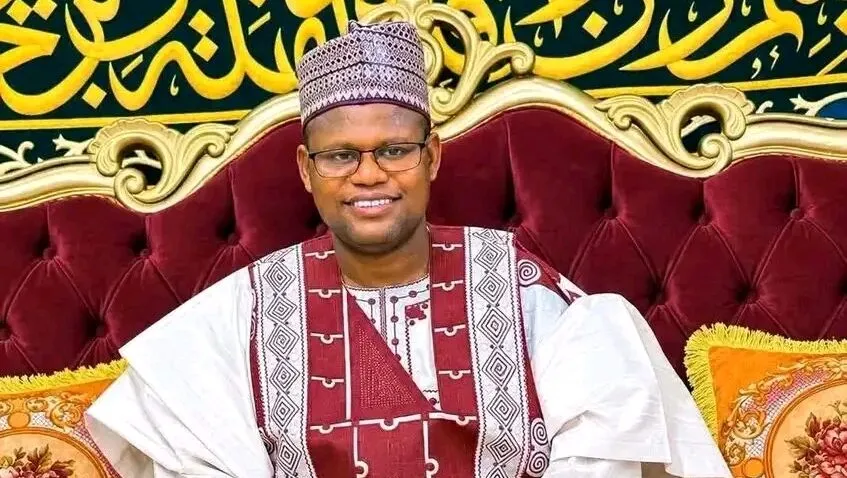Sheikh Yahaya Masussuka, the Katsina cleric, in a white and maroon traditional outfit and glasses, smiling on a red sofa