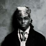 FOLA in black and white, wearing a patterned bandana, tinted glasses, a dark coat and a diamond chain