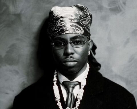 FOLA in black and white, wearing a patterned bandana, tinted glasses, a dark coat and a diamond chain