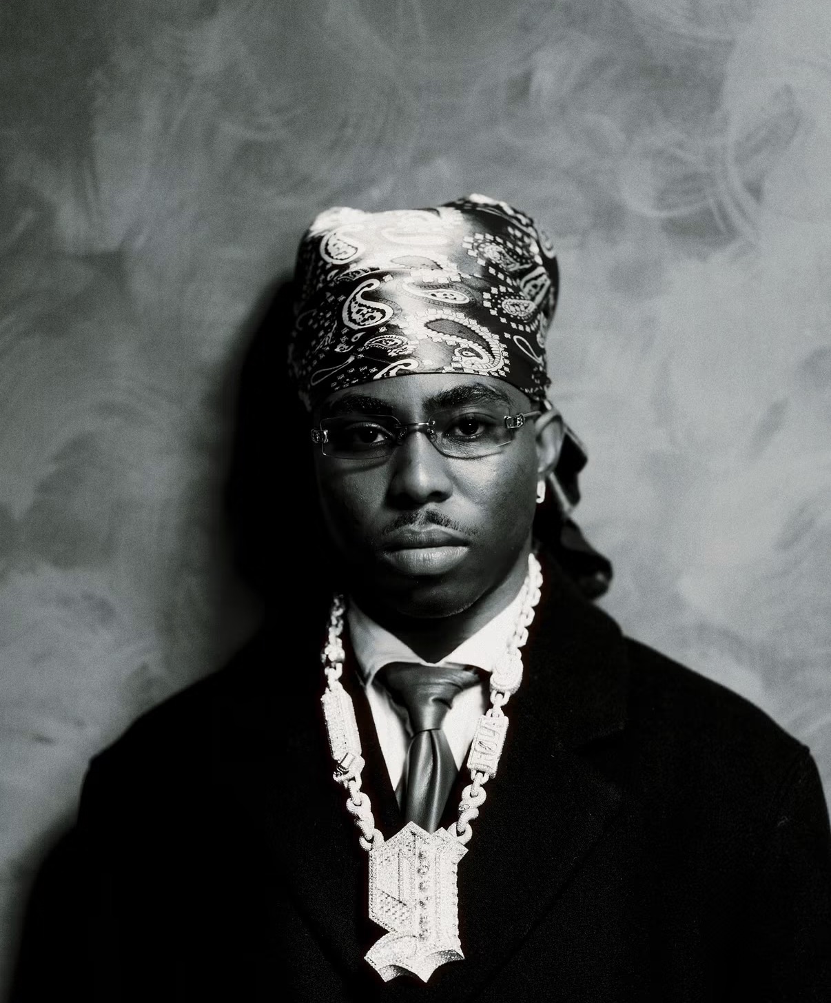 FOLA in black and white, wearing a patterned bandana, tinted glasses, a dark coat and a diamond chain