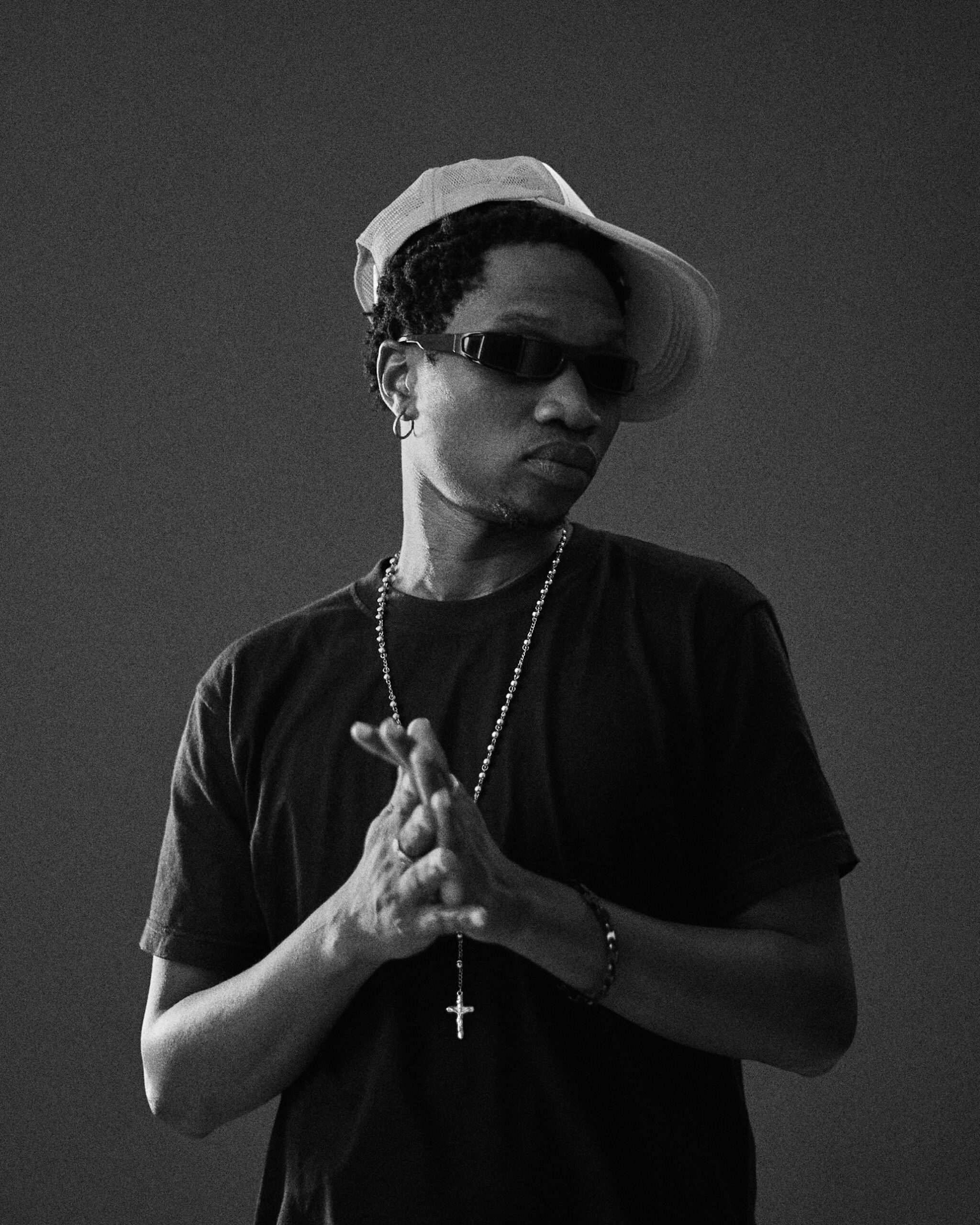 Black-and-white portrait of a man in a trucker cap and sunglasses with a rosary chain, hands pressed together