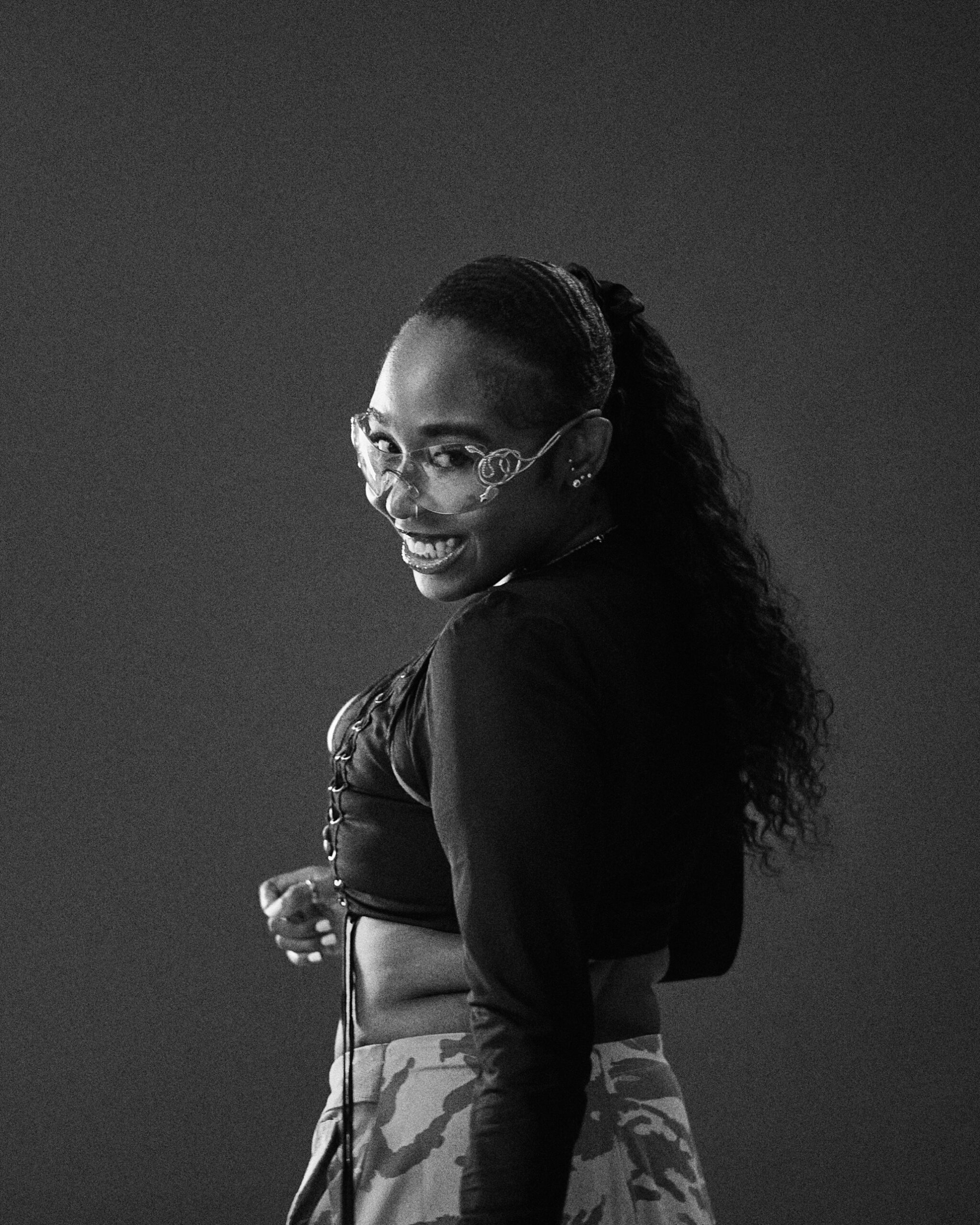 Black-and-white portrait of a woman in clear sculpted glasses looking back over her shoulder with a wide smile