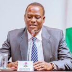 A Nigerian government official speaking at a table with a Head of Delegation name card and the Nigerian flag behind him