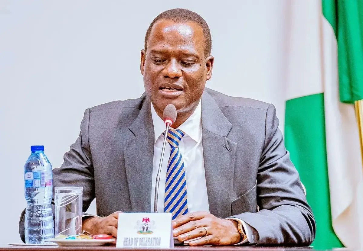 A Nigerian government official speaking at a table with a Head of Delegation name card and the Nigerian flag behind him