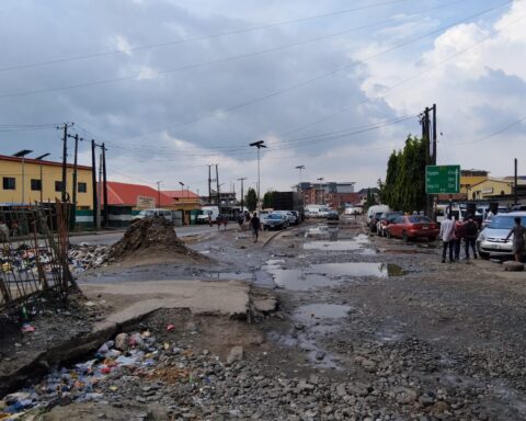 A flooded, broken road in Lagos with potholes, piled-up rubbish and cars parked along the side