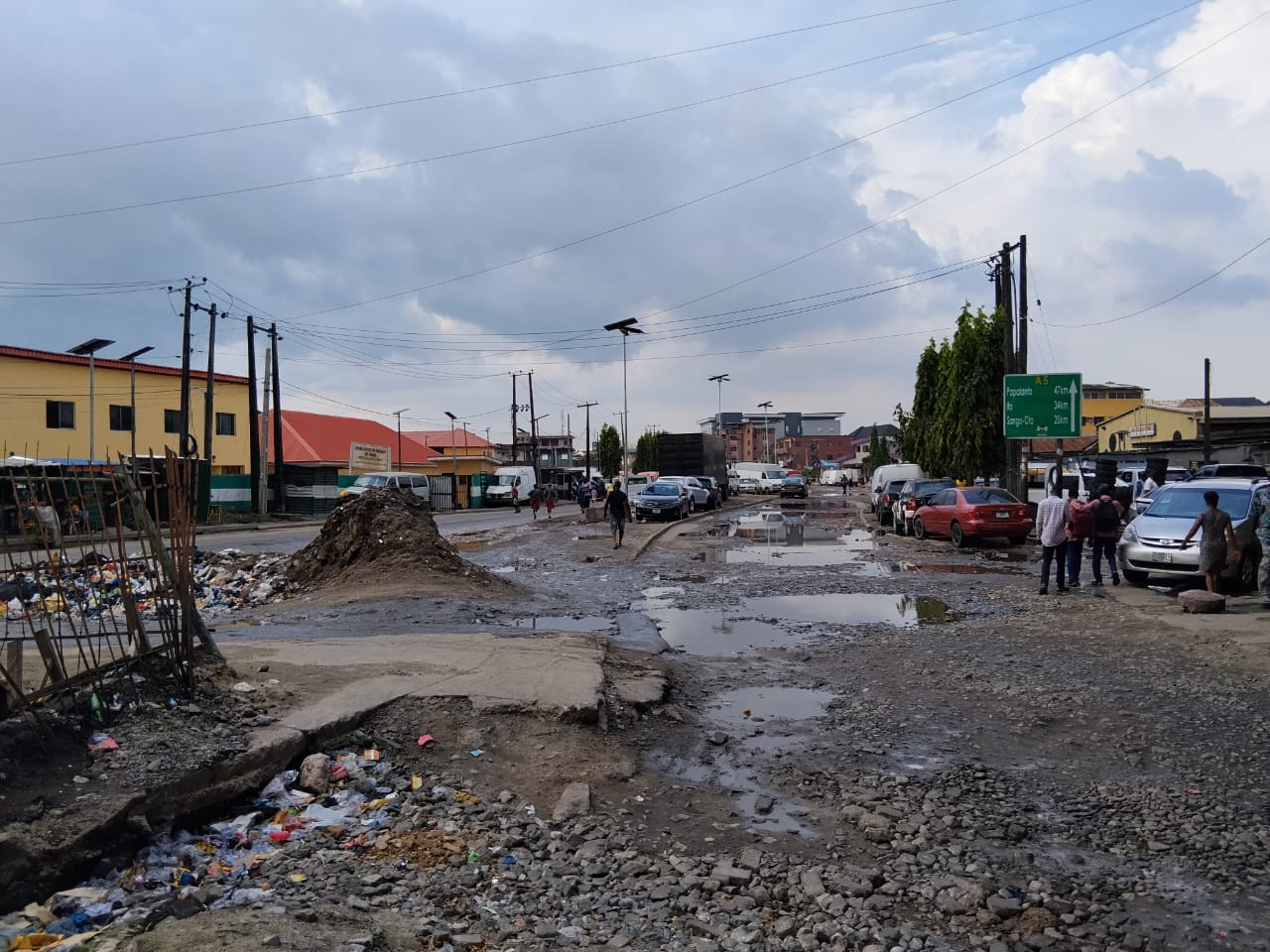 A flooded, broken road in Lagos with potholes, piled-up rubbish and cars parked along the side